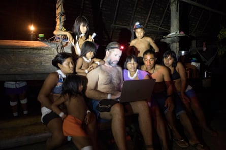 Bruno Pereira sits surrounded by a group of Indigenous men, women and children, lit up by the light of his head torch. He is showing them footage on his laptop