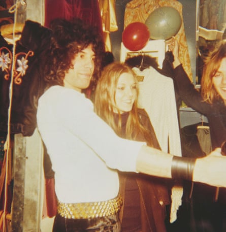 A dark haired man in a white top and black trousers with a blond woman at a party with balloons behind them