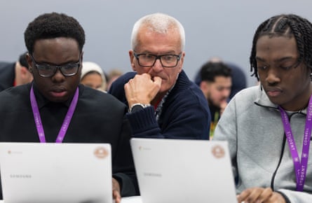 The Hacking Games designer John Madelin looking over the shoulders of two young men working on laptops.