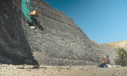 A sheer cliff face, with excavation equipment suspended partway up and three workers in helmets looking up from the ground