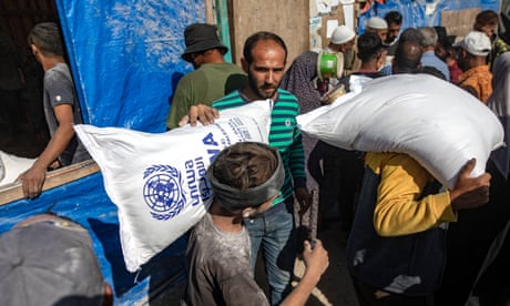 A man handing a white sack containing flour to a boy in a grey t shirt, who is balancing it on his shoulder