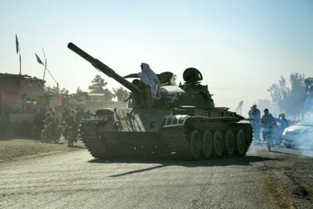 a fighter in white robes and headscarf sits behind a huge gun on the tank, silhouetted against the sky. The road looks dry and dusty, with low-rise buildings in the background.