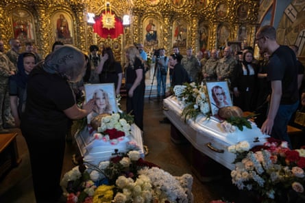 Mourners gather around two white coffins in gilded surroundings. Each coffin has sitting on it a portrait of the deceased