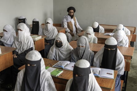 Cowed-looking girls in full hijab and veils sit in rows of desks at school with a bearded man in a turban sitting at the back of the room 