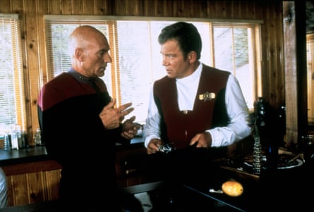 Captain Picard and Captain Kirk stand talking intently indoors near a window
