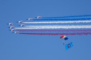 A parachutist with a French tricolour chute descends holding a large flag and jets fly past in formation in the background leaving red, white and blue vapour trails behind them