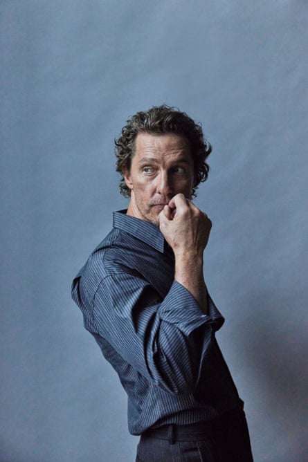 Portrait of Matthew McConaughey shot from the side with his hand to his mouth is smart dark blue shirt and dark trousers.