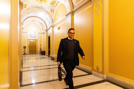 Tall man with dark hair and glasses and dark suit walks through ornate lighted round-ceiling hallway.