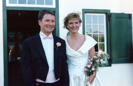 A man in black dinner jacket white shirt and pink bow tie with a woman in a white wedding dress with wide low necked collar, and holding pink flowers