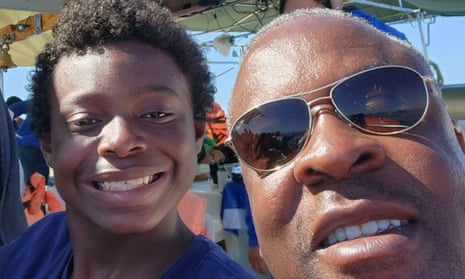 A selfie taken in the son of a Black teenager with curly black hair and a Black man wearing sunglasses, and a bald head, both smiling, maybe at an outdoor festival or a theme park.