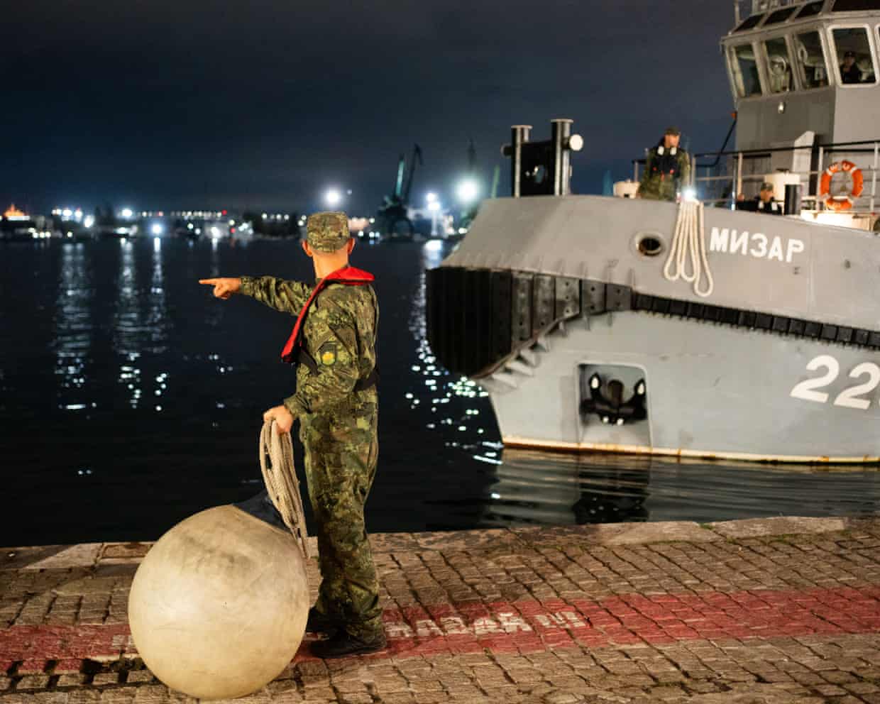 A Bulgarian navy sailor points while holding rope, with the Mizar rescue tug moored nearby at night