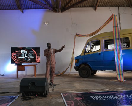 A man stands in the corner of a large concrete-floored hall, speaking into a microphone. Behind him is a TV on a table, which is showing the logo of the biennale and next to him is a swag of rainbow-coloured material hanging from the ceiling and a blue and white van.