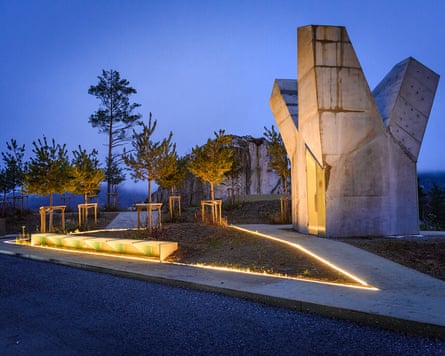 A sculptural concrete monument with angular peaks stands illuminated at dusk, beside a lit pathway and young trees.