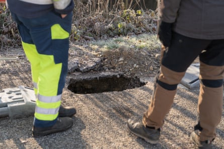 Two pairs of legs standing next to a hole in a tarmacked surface.