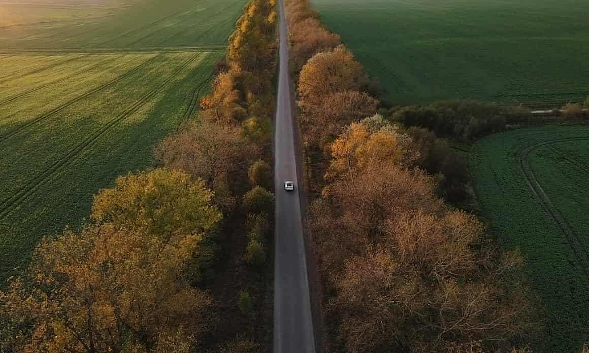 An aerial view of an electric car driving on a country road at sunset, passing golden trees and green fields.