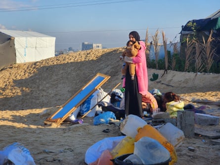 Palestinian woman holding a child as she stands alongside their possessions