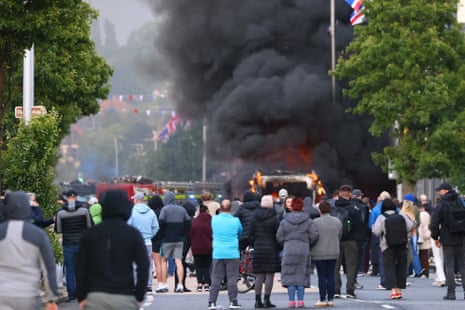 Pessoas assistem enquanto um veículo pega fogo durante um protesto após um incidente de esfaqueamento no norte de Belfast.