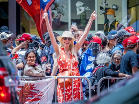 Among a crowd, a tall middle-aged white woman with long blond hair, sunhat, and red dress grins and holds both thumbs up.