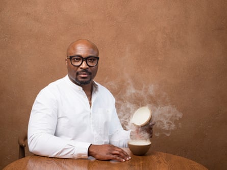 Aji Akokomi is wearing a white shirt and black glasses. He sits at a round table and is lifting the lid from a steaming bowl