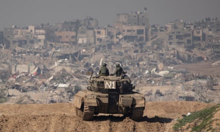 Two soldiers standing on top of a tank on a ridge overlooking a landscape of destroyed buildings