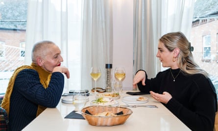 An older woman with very short cropped grey hair and bright red lipstick, wearing a mustard sleeveless polo neck over a black long-sleeve top, and a younger woman with long blonde hair pulled back from her face, wearing a black polo neck with a single pearl necklace, sitting chatting at a restaurant table