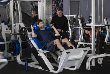 a man stands by as another man uses a piece of exercise equipment in gym