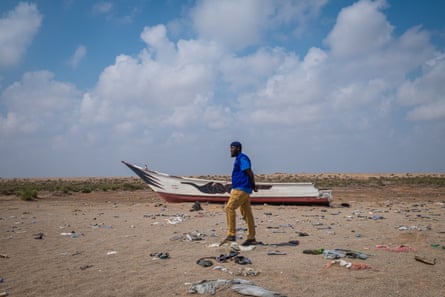 A man walks along a beach strewn with clothes and debris. A wrecked boat is in the background.