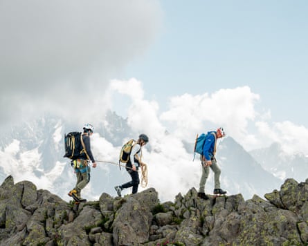 Fully equipped mountaineers walking over a rocky ridge against a backdrop of snowy mountains.