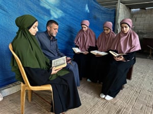 A man and four women sit indoors reading the Qur’an. The women wear long black dresses with headscarves, one in green and the others in pink, while the man wears a dark grey outfit.
