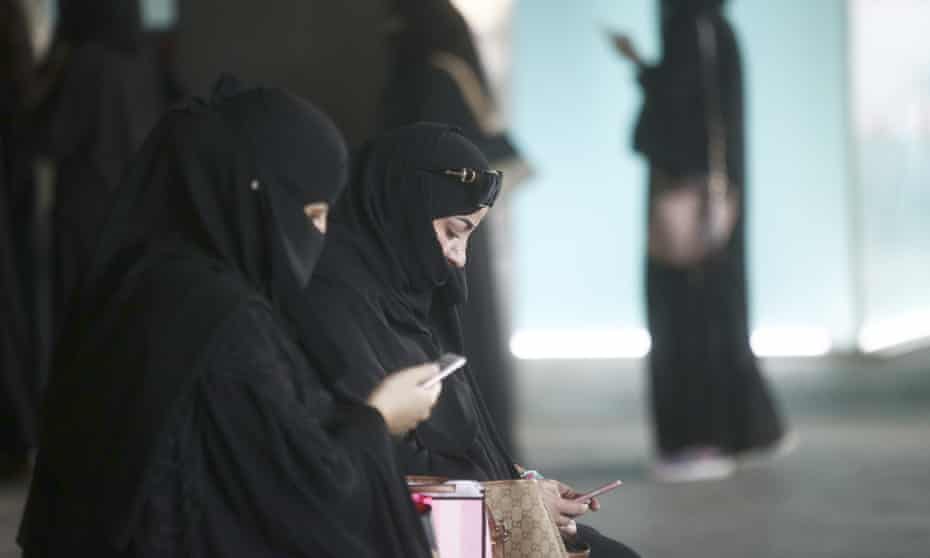 Female shoppers wearing traditional Saudi Arabian dress check their smartphones whilst waiting for transport outside the mall.