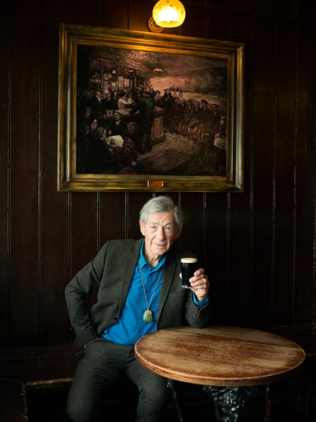 Sir Ian wearing a black suit and blue shirt sits underneath an old gilt-edged painting and raises a glass of Guinness at The Grapes pub in 2012