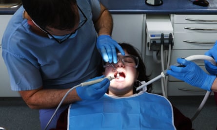 A patient is treated at a NHS dental clinic in London: her open mouth is spotlit in the centre of the photo as a dentist holds instruments to it; the hands of a dental nurse or assistant are also seen; the patient has a blue bib on, the dentist has blue scrubs and blue rubber gloves, and the assistant holding a suction tube also wears blue rubber gloves.