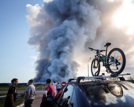 People stand on a roadside watching a large plume of dark smoke rising in the distance, with a car and bicycle visible in the foreground