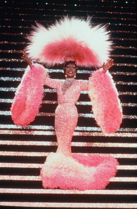 A woman wearing an extravagant sparkly pink dress with huge fur trim to the extremities and a huge fur headpiece at the bottom of a sparkly staircase