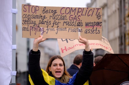 A woman holds a placard that reads 'Stop being complicit consuming Russian gas, selling parts for Russian weapons and profiting from frozen Russian assets'