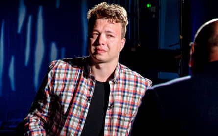 A man in a checked shirt looks at another man, seen in silhouette, during a TV interview