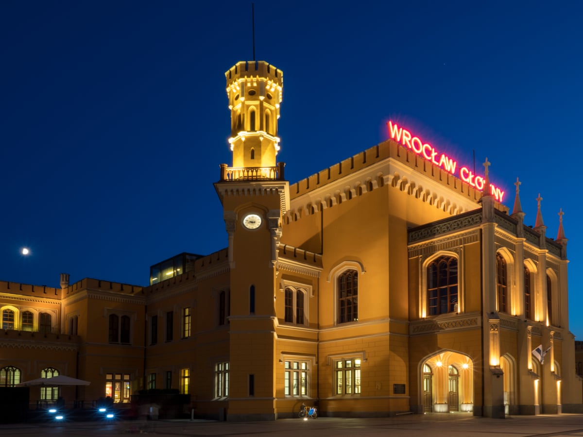 A yellow neogothic railway station at night, topped with red neon letters saying Wrocław Główny
