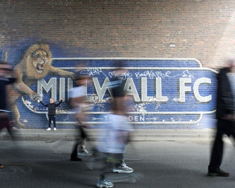 A fan poses next to a mural outside the Den whilst other fans walk past.