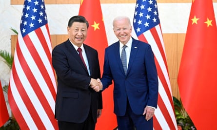 Xi Jinping and Joe Biden are shaking hands and smiling as they stand in front of two Chinese and two US flags