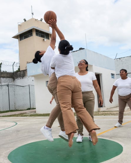 Two women, one barefoot, jump for a basketball on an outdoor court as others look on