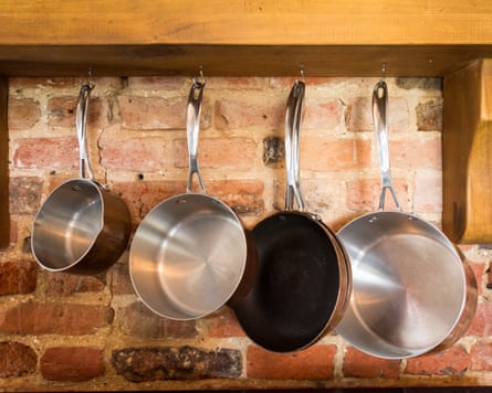 Metal saucepans hanging from a large chunky wooden shelf against a bare brick wall