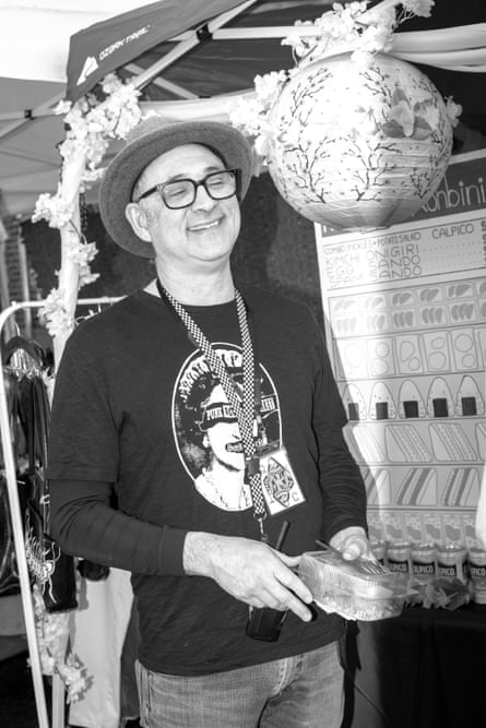 A black-and-white image of a smiling man in a fedora, hat, black long-sleeve T-shirt, jeans, and his phone on a cord around his neck.
