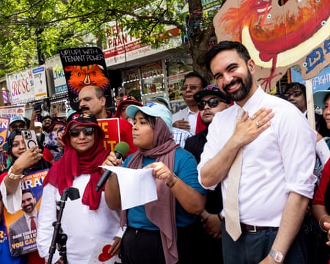 A man holds his hand to chest and smiles while standing next to a woman talking into a microphone as a crowd of protesters watch on