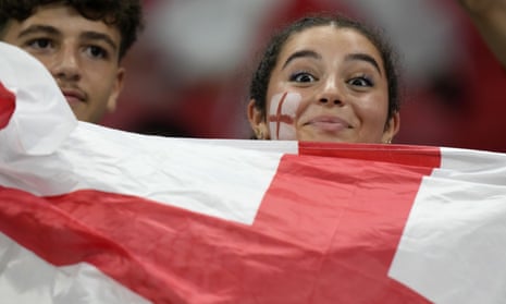 A fan holding an England flag smiles at the camera from the stands while waiting