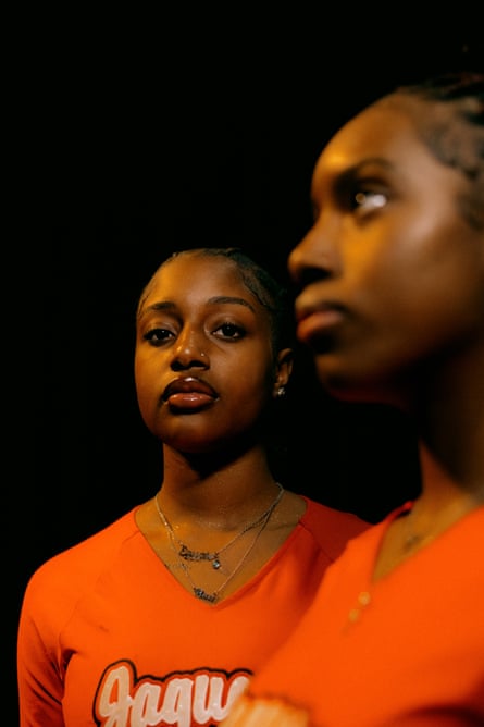 A portrait of two Black teenage girls, both in bright orange shirts that say “Jaguar.” The girl on the left, in the background, is in focus and looks seriously at the camera. The girl on the right, in the foreground, is blurred and looks in front of her, to our left.