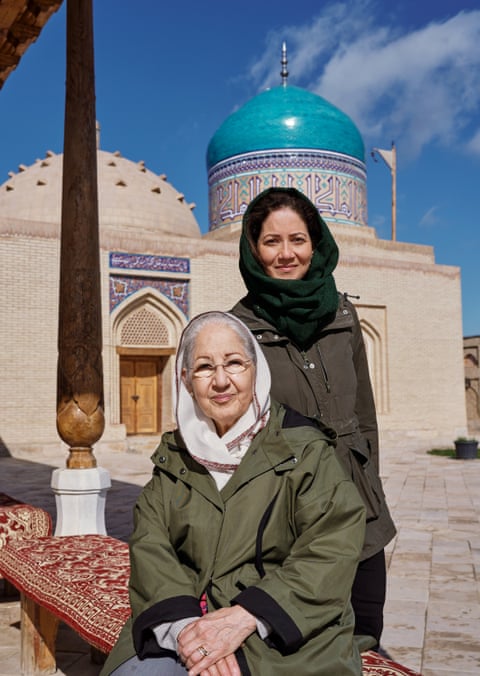 Mishal Husain, wearing a green headscarf and coat, and standing next to her mother, Shama, who is also wearing a coat and headscarf, and sitting on an upholstered bench outside a domed and decorated mausoleum