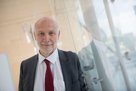 Peter Fonagy stands next to a glass door which reflects him a little. He has a short white beard and bald head, and is smiling.