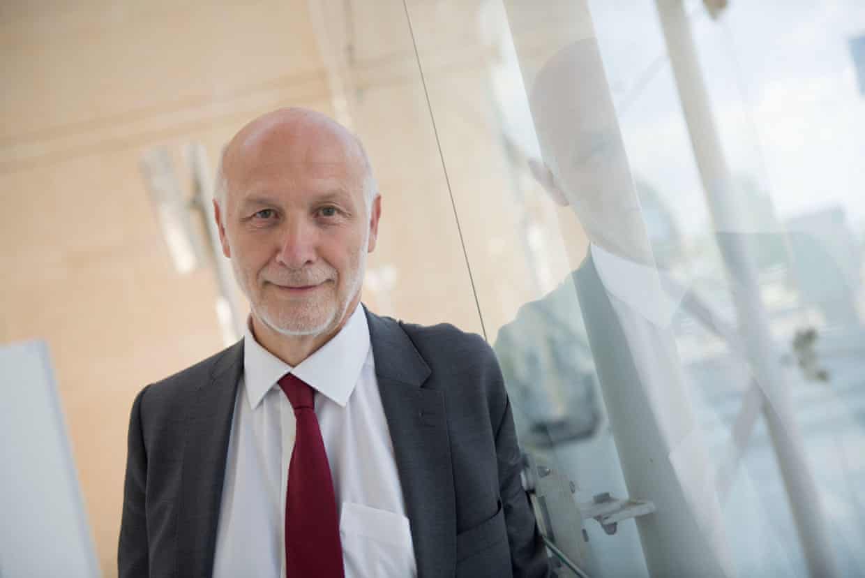 Peter Fonagy stands next to a glass door which reflects him a little. He has a short white beard and bald head, and is smiling.