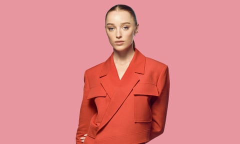 Head shot of actor Phoebe Dynevor in orange jacket against pink background