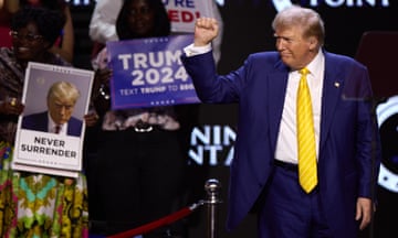 Man in suit and yellow tie raises hand on stage with supporters' signs behind him